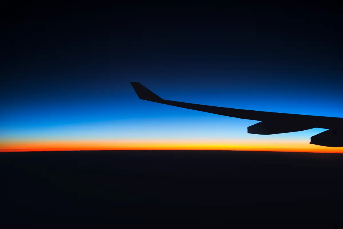 An aircraft wing above the clouds with a band of orange sunrise on the horizon